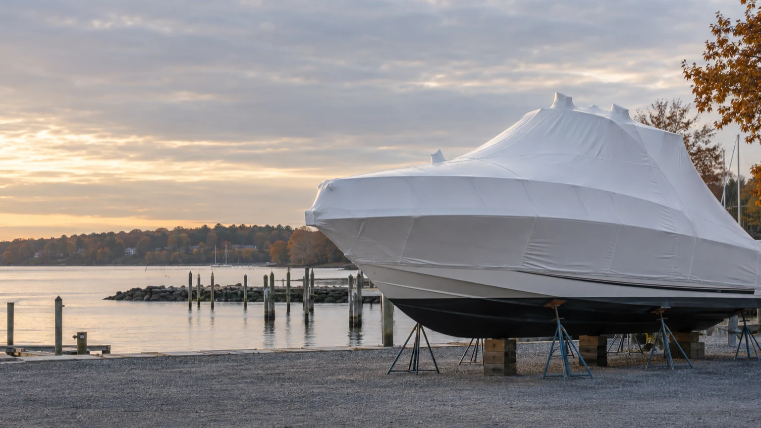 A white shrink-wrapped powerboat stored on stands beside a marina in autumn